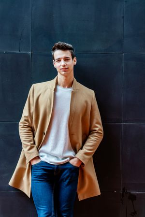 Man leaning on dark wall outside and wearing a camel coat while looking at camera