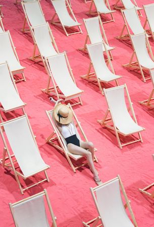 Woman in bathing suit wearing a sun hat lying on one of the arranged beach lounger on pink surface
