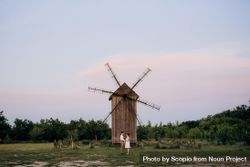 Man and woman holding hands and standing beside wooden windmill on green grass field bx82jb