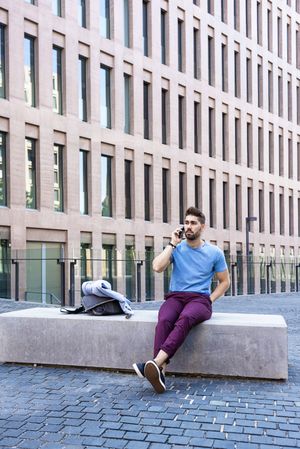 Portrait of man in blue t-shirt speaking on phone while sitting outside building
