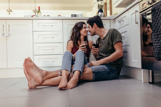Young man and woman sitting on kitchen floor holding wine glasses