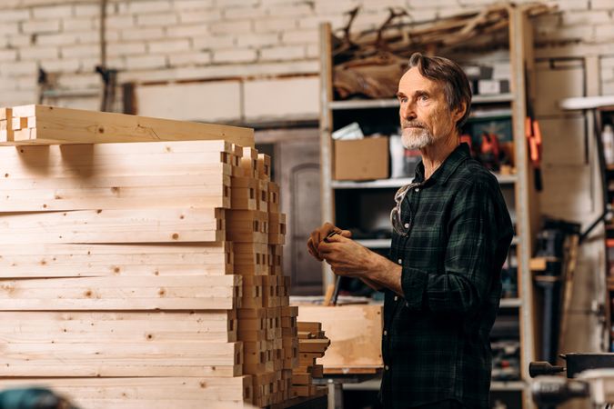 Older male carpenter next to pile of wooden planks in shop