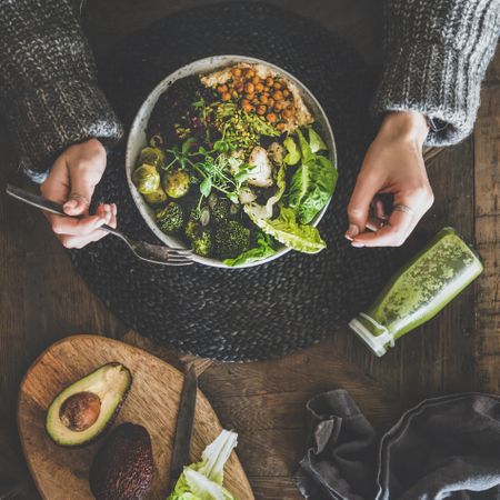 Healthy vegetarian bowl pictured on table with smoothie on side and hands with fork, square crop