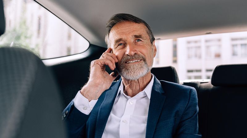 Smiling mature man with grey beard taking a call in backseat of car
