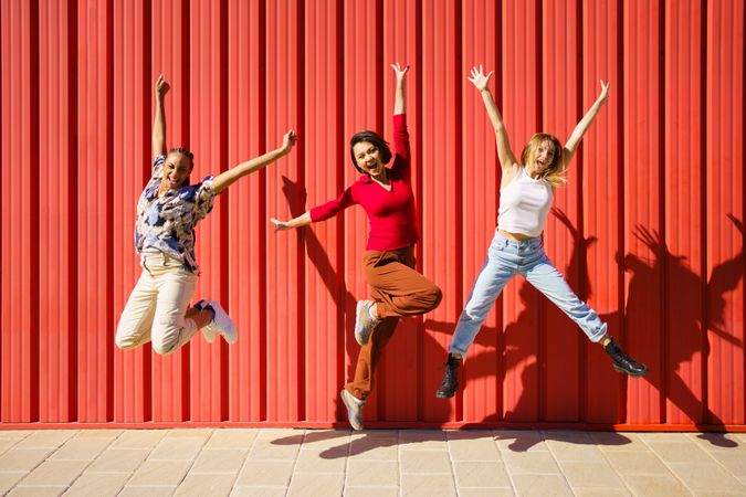 Three women jumping for joy in front of red wall on sunny day