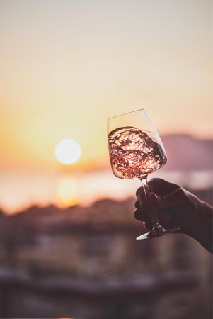 Man's hand swirling glass of rose wine against sunset, vertical composition