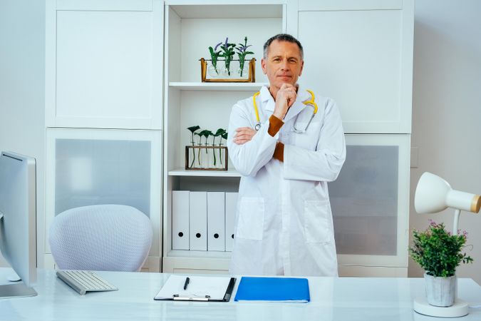 Contemplative mature doctor standing in a clinical setting with hand to his chin