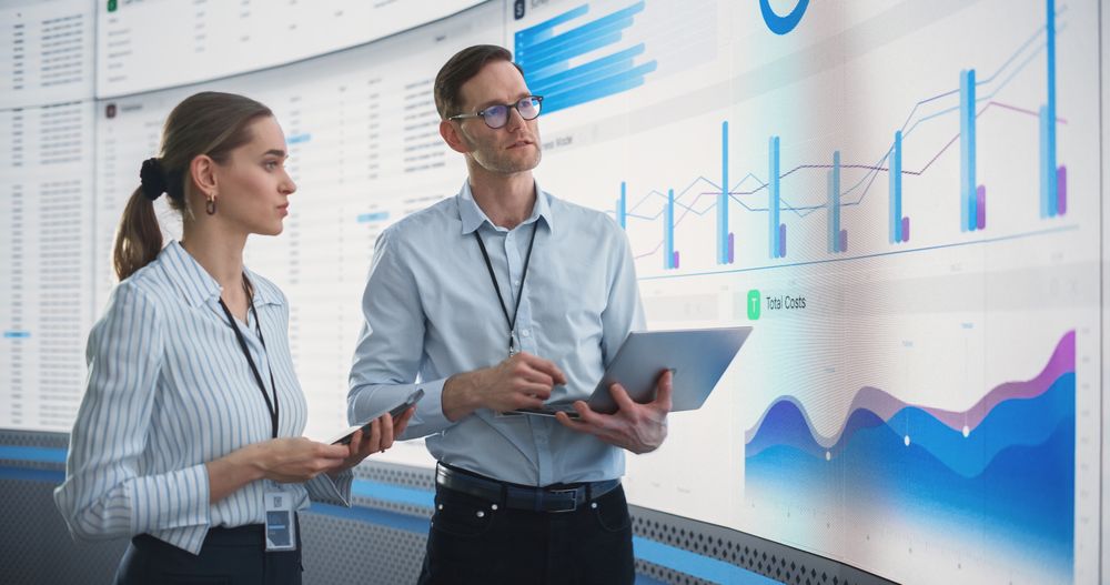 Male And Female Data Analysts Discussing New Locations For Business In Front Of Big Digital Screen With City Map In Monitoring Office.