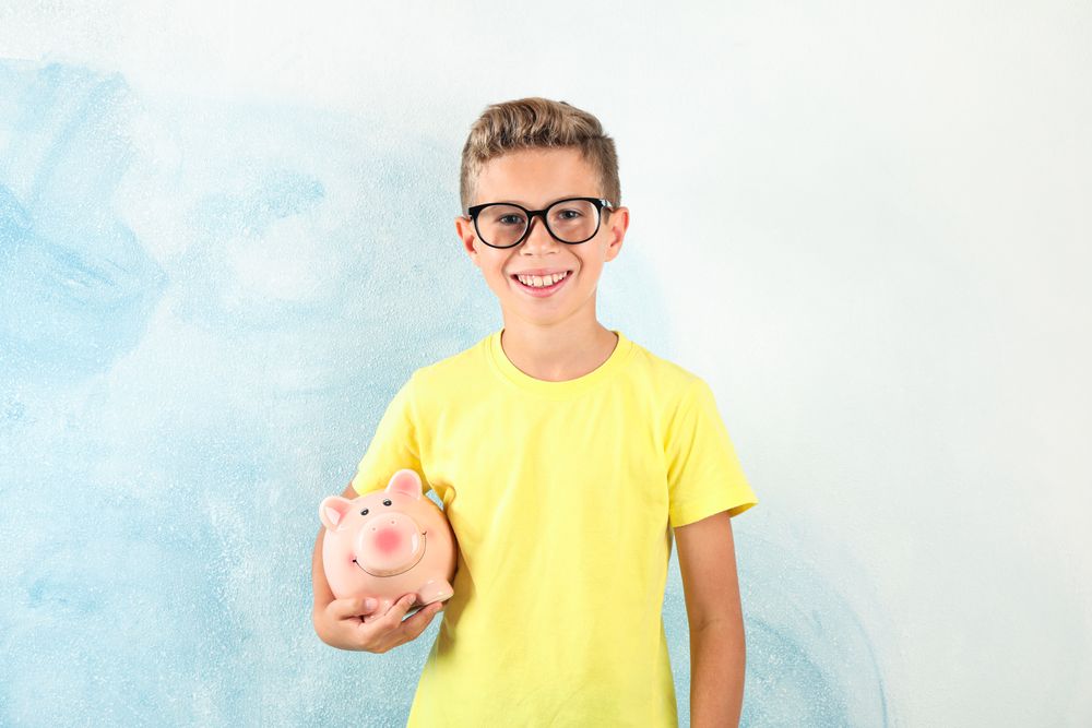 Boy in glasses with piggy bank against color background, space for text