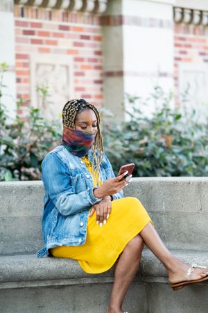 Woman wearing mask sitting in front of library reading messages on mobile phone