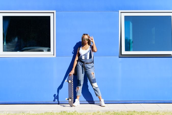 Female in denim overalls adjusting sunglasses in front of blue building
