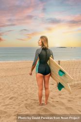 Woman standing on beach with surfboard gazing out at beautiful view, vertical composition 4Njz24