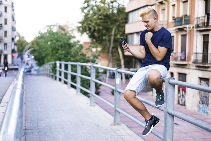 Young male with blonde bangs sitting on railing while using a mobile phone