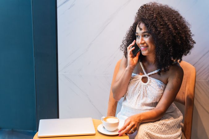 Smiling Black woman speaking on smartphone with cup of latte