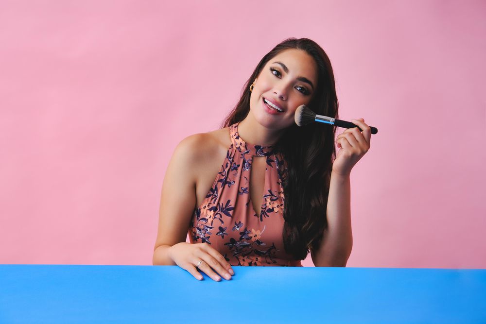 Smiling woman with long brown hair holding large make up brush to her face
