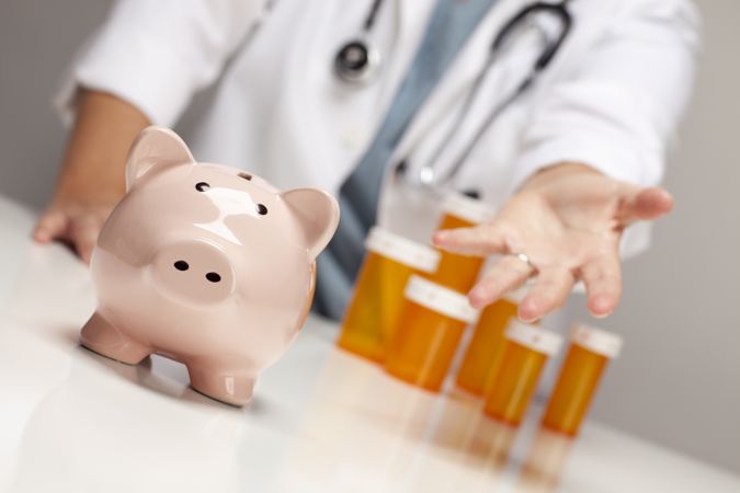 Doctor Reaches Palm Out Behind Medicine Bottles and Piggy Bank