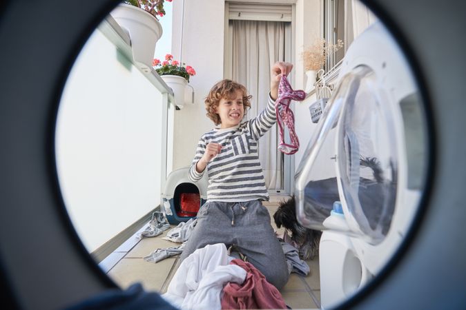 Boy sitting near washing machine and holding panties with disgus