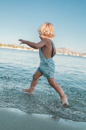 Fair haired cute toddler running in calm water of sea on shore