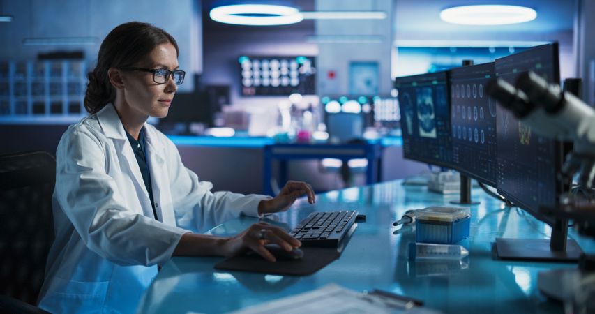 Medical Research Center Laboratory: Female Neurologist Using Desktop Computer With MRI And PET Scans Of a Brain.