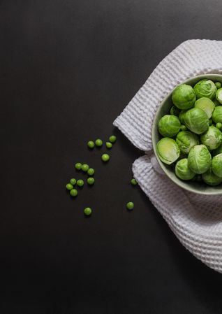 Bowl containing brussels sprouts on dark background with some green peas and a towel  on tabl