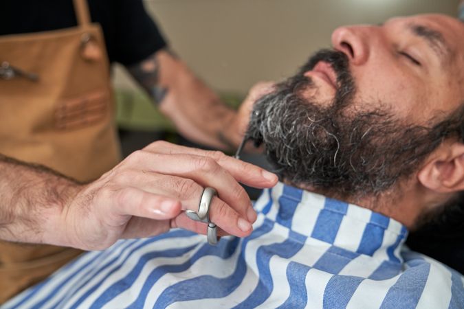 Crop hairdresser cutting beard of man in barbershop