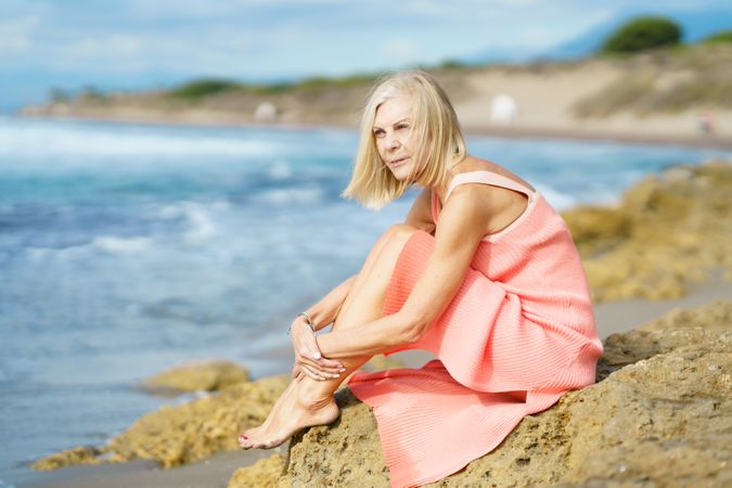 Serene older female with grey hair sitting on coastal rocks enjoying the view