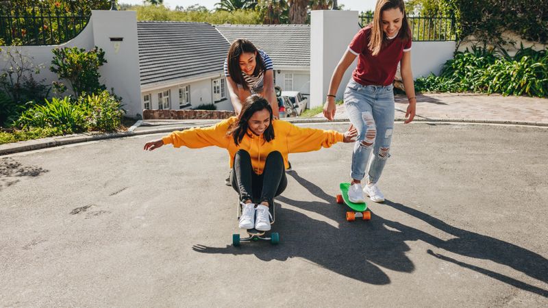 Smiling girl pushing her friend who is sitting on a longboard from behind