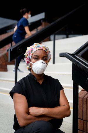 Portrait of female nurse in scrub hat and mask sitting outside on steps