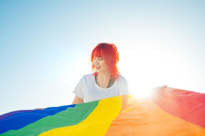 Young woman with red hair holding rainbow flag under blue sky