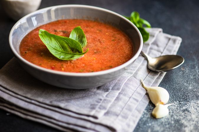 Grey bowl of gazpacho soup with basil leaves and garlic on napkin