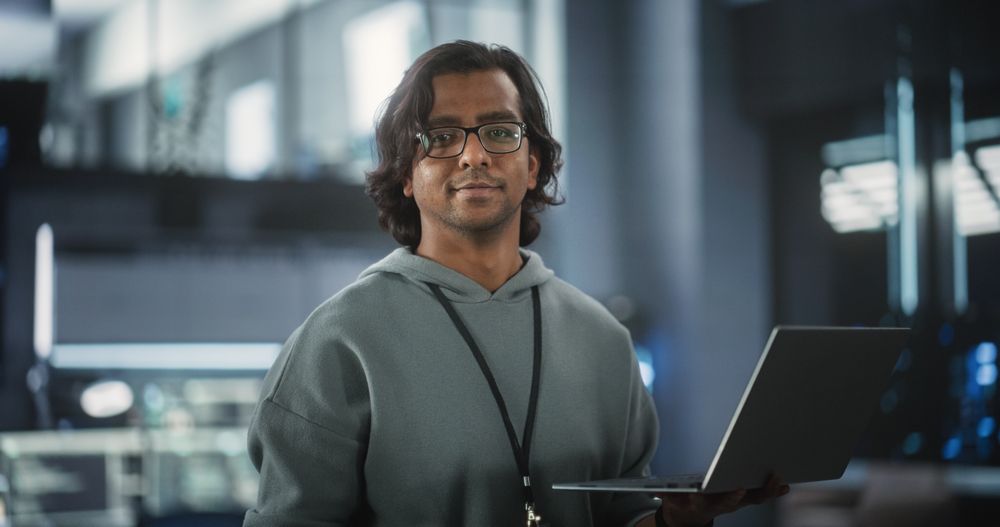Young Indian Software Engineer Using Laptop Computer in Data Center, Looking at Camera.