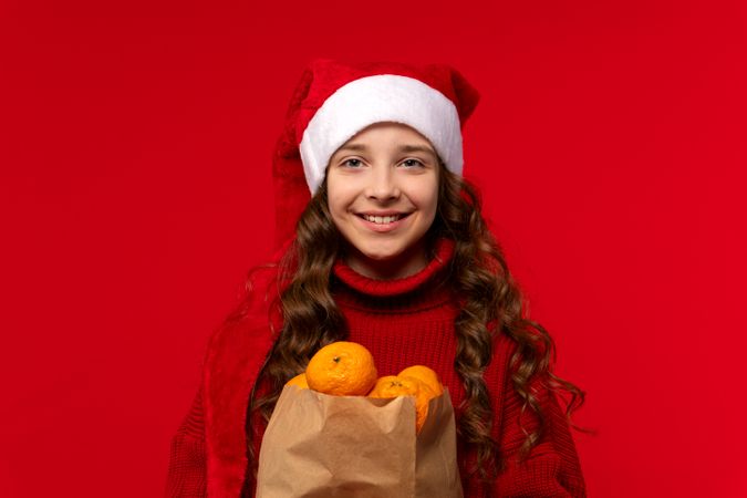 Smiling girl in Santa hat holds bag filled with oranges.