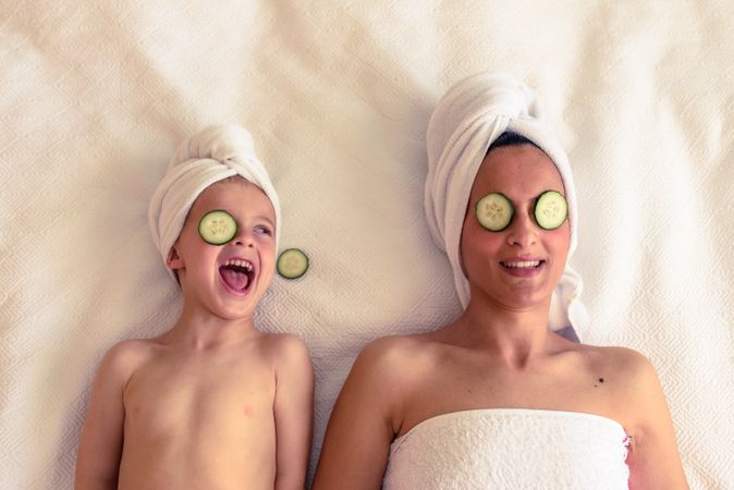 Cheerful kid with mother with cucumber on eyes