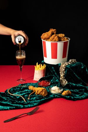 Woman pouring soda in wineglass beside collections of jewelry and fast food