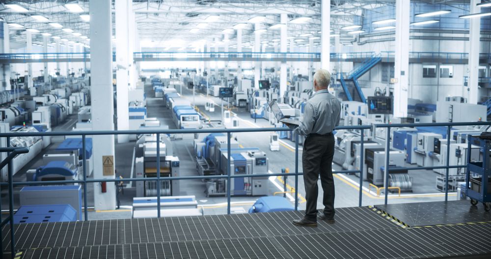 Factory Manager Working on Laptop at a Futuristic Industrial Manufacture with Autonomous AI Robots. Specialist Monitoring All the Data on a Computer, Checking Reports and Planning Workload