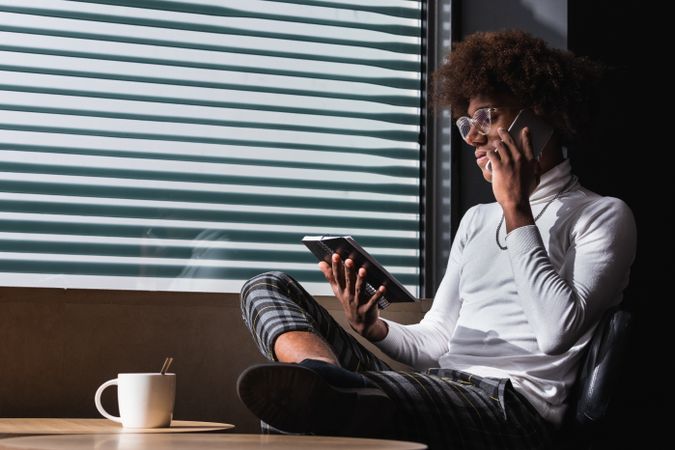 Fashionable young Black male journalist writing article and talking smartphone in cafe