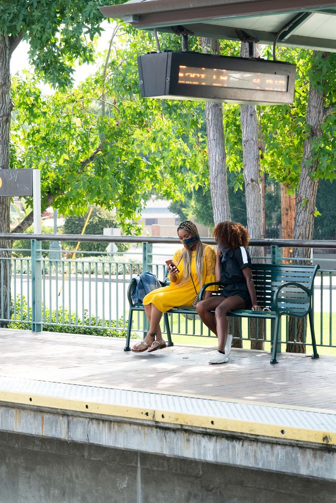 Two women sit outdoors on bench on train platform looking at schedule on mobile phone