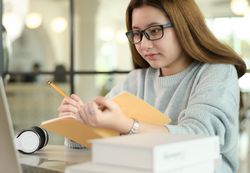 Young woman with a pen and a notebook is taking notes from laptop