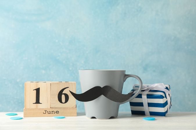 Cup with decorative mustache, gift box and wooden calendar on table against blue background, space for text