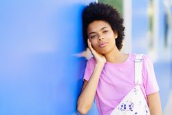 Serious woman looking at camera while holding her face and leaning her side on blue building
