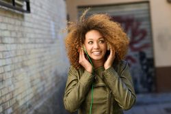 Happy woman with curly hair and green jacket pictured using headphones in backstreet