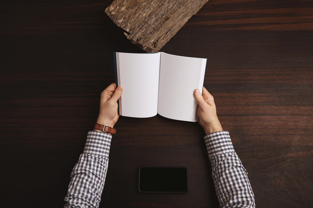 Arms in plaid shirt holding open book with cell phone on table