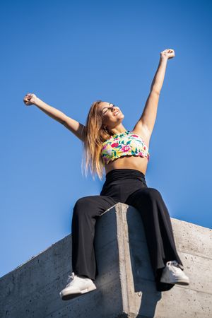 Happy woman sitting on corner of roof with arms up, vertical