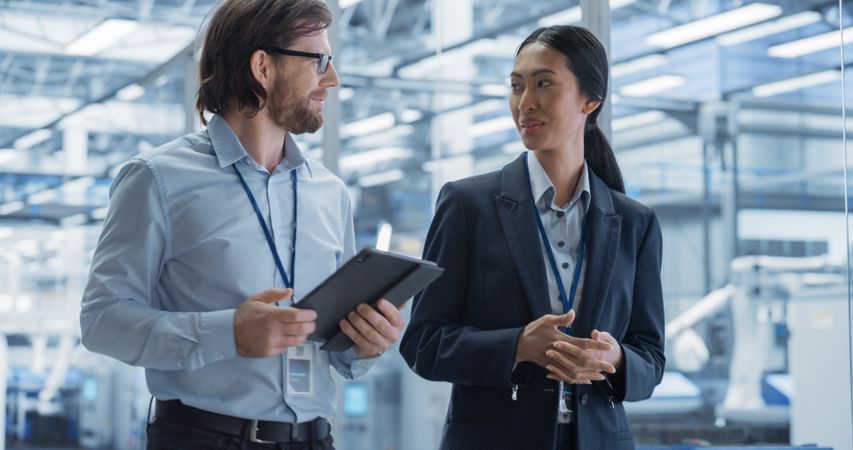 Two Diverse Engineers Walking in Office, Using Tablet Computer at a Modern Electronics Factory. Female and Male Specialists Talking, Monitoring Robots on Modern Assembly Line.