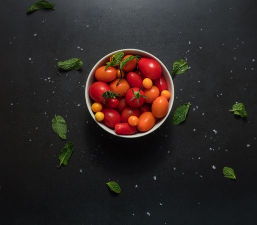 Bowl containing tomatoes placed on a dark background with mint scattered around