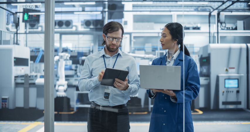 Engineer and Asian Female Project Manager Standing in Office, Talking, Using Computers and Monitoring Production at a Modern Automated Electronics Manufacture with AI Robotic Arms