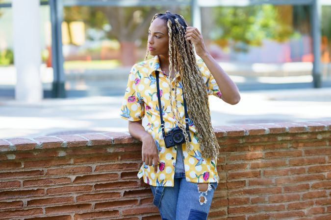 Female in bold patterned shirt standing next to brick wall with camera