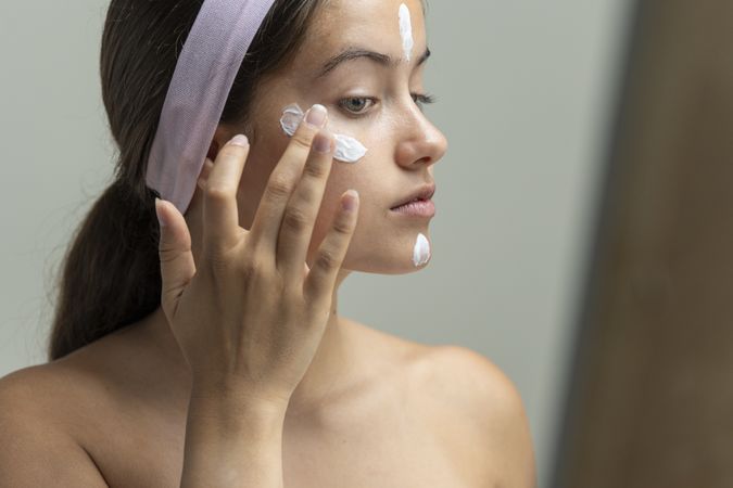 Young woman looking in the mirror, preparing her skin with cream for a beauty routine