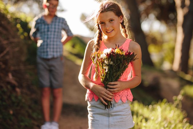 Smiling girl with a bouquet of flowers in hand standing in park with a boy in the background