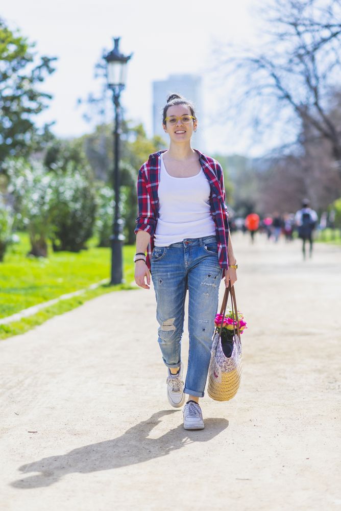 Front view of a young smiling hipster woman walking in a park in sunny day while holding a wicked basket